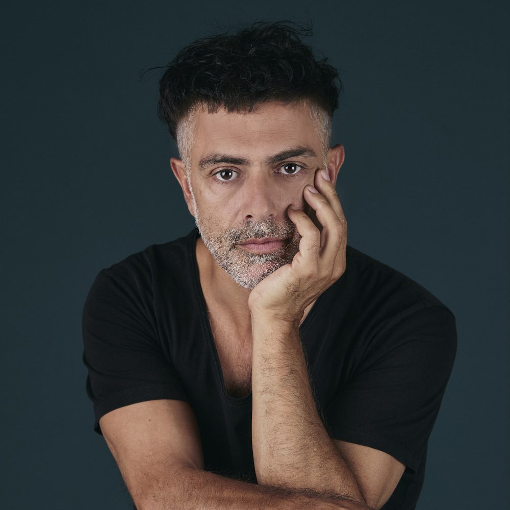 2832053 Portrait en plan serré d’un homme dans la cinquantaine, cadré de face, sur fond gris foncé uni. Il a les cheveux courts, poivre et sel, légèrement ébouriffés sur le dessus. Son visage est allongé, avec des sourcils foncés, des yeux bruns regardant droit vers l’objectif, une barbe courte et soignée, également grisonnante. Son expression est neutre, sérieuse, presque introspective. Il porte un sweat à capuche noir, sans motif visible. La lumière est douce et frontale, mettant en valeur les traits de son visage et créant un léger modelé sur les joues et sous le menton. L’ensemble donne une impression sobre, intime et posée.