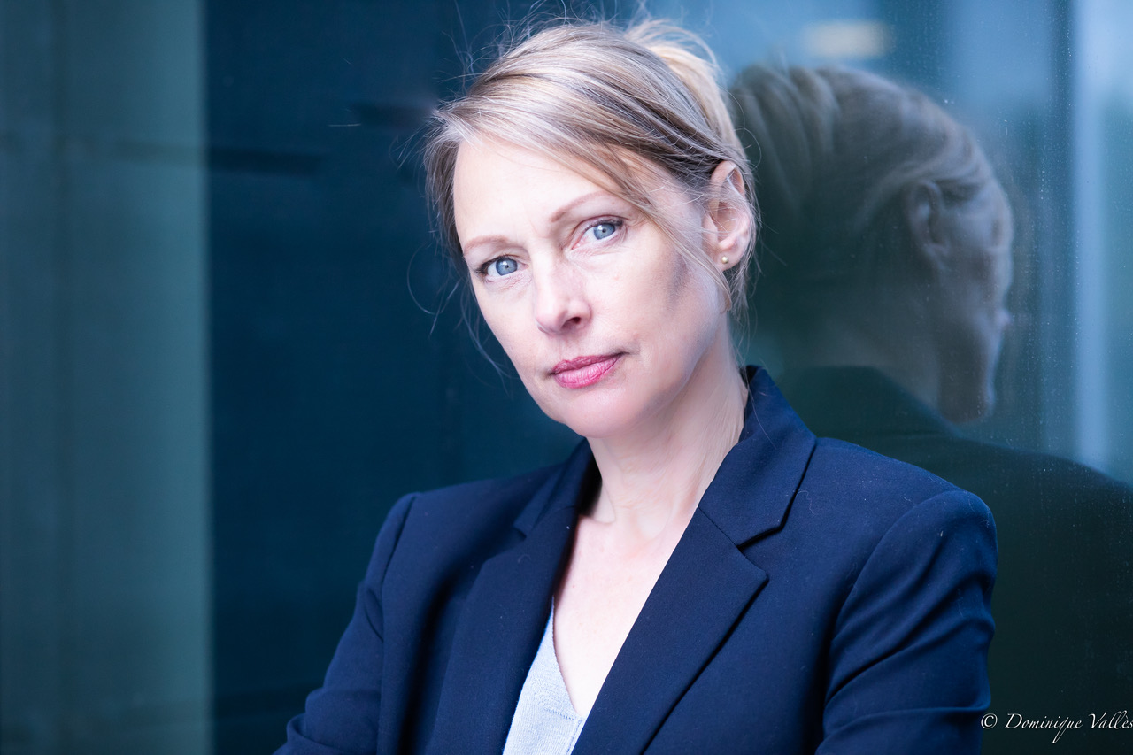 alexandra-tiedemann_127a5590 En plan rapproché, une femme, la petite cinquantaine se tient près d’une paroi vitrée. Elle a les cheveux blonds clairs, attachés en arrière, laissant apparaître un visage aux traits fins et aux yeux bleus très clairs. Son regard est direct, intense, légèrement interrogateur. Elle porte une veste sombre, structurée, sur un haut clair. La lumière est froide, presque bleutée, et se reflète dans la vitre à côté d’elle, où l’on distingue son reflet. L’ensemble dégage une atmosphère sobre, élégante et contemporaine.