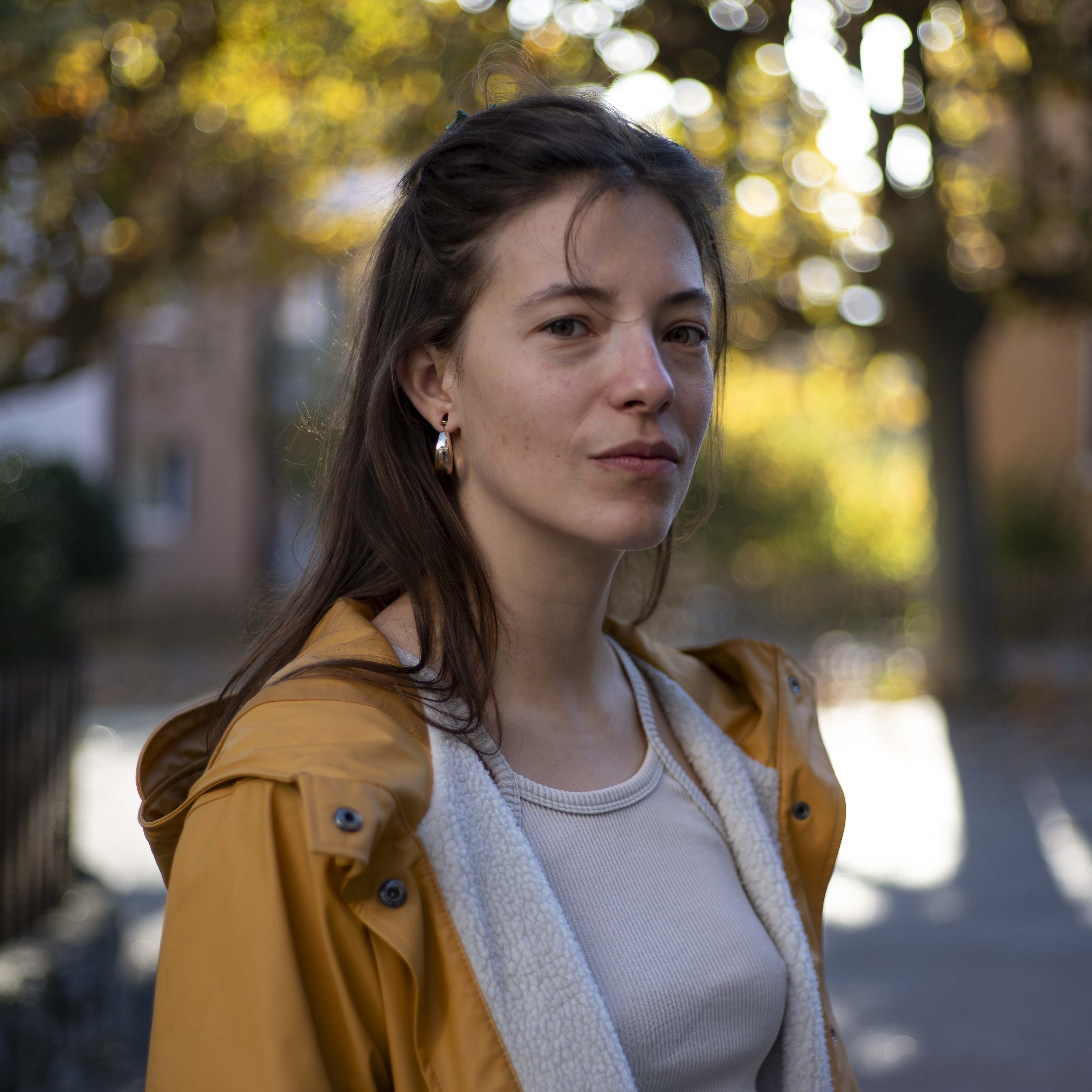 BaladeFall-Salma_10 (1) Une jeune femme la mi-vingtaine se tient face à l’objectif. Elle a les cheveux bruns, mi-longs, légèrement ramenés en arrière, et porte de petites boucles d’oreilles dorées. Son visage est fin, au teint clair, avec une expression calme, presque méditative. Elle porte un imperméable jaune moutarde, ouvert sur un haut clair près du corps, doublé d’une matière douce et écrue visible à l’intérieur. L’arrière-plan est flou : des taches lumineuses de vert et d’or filtrent à travers les arbres, et l’on devine des façades de maisons. La lumière est douce, chaleureuse, typique d’un après-midi d’automne.