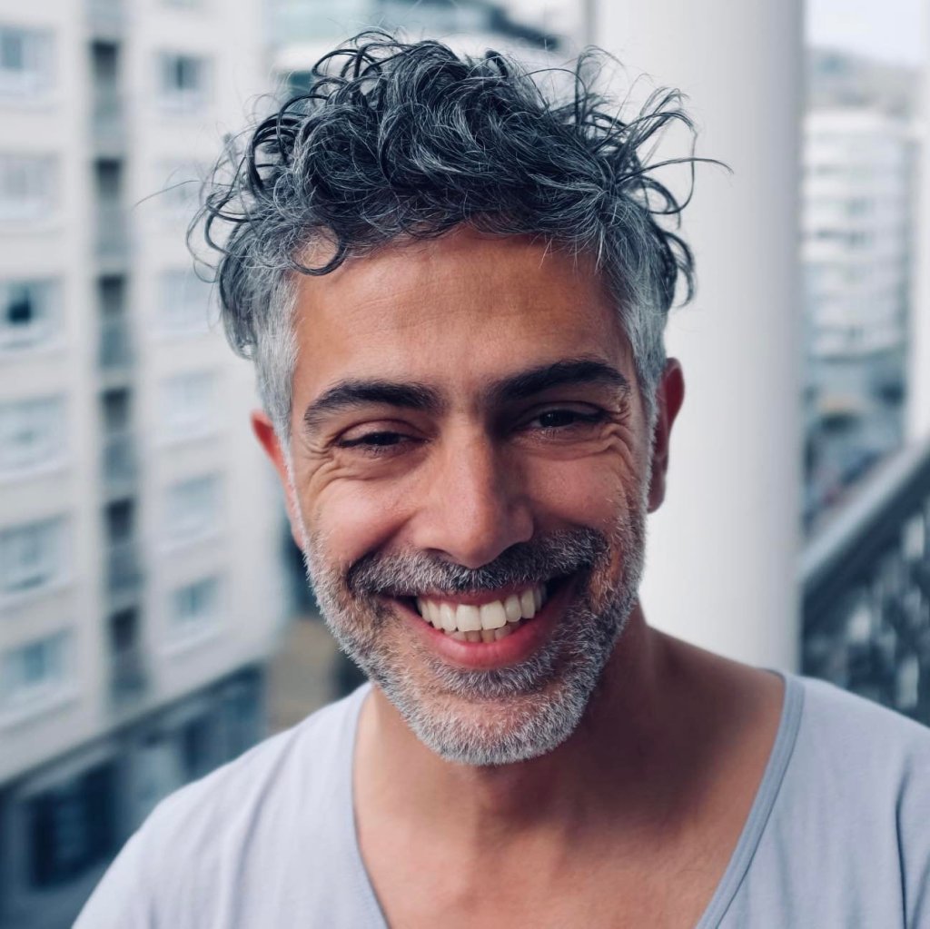 En plan rapproché, un homme la cinquantaine fait face à l’objectif. Il a les cheveux courts, bouclés, poivre et sel, un peu ébouriffés, et une barbe courte de la même couleur. Son visage est éclairé par un large sourire qui découvre ses dents et creuse légèrement les rides autour de ses yeux.
Il porte un t-shirt gris clair. Derrière lui, l’arrière-plan est flou : on devine des immeubles clairs et un balcon, dans une lumière douce et légèrement froide qui évoque un décor urbain.